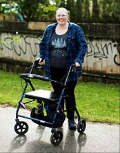 A photo of Heather smiling with their walker against a graffitied wall next to patch of grass by the cement pavement. They wear a blue and black plaid shirt with a t-shirt underneath that reads “Forest Friend” in white text alongside a graphic of trees.