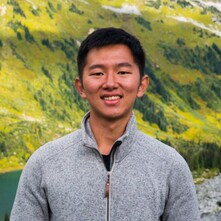 A torso and up picture of WeiChun wearing a grey half-zip sweater. The background is a green slope with trees on it. 